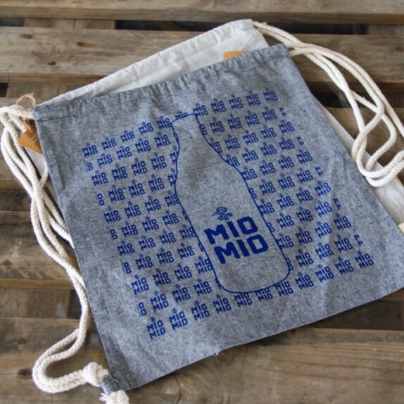 Dunkelgrauer Mio Mio Baumwollbeutel mit blauem Logo und weißen Bändern auf Holzboden