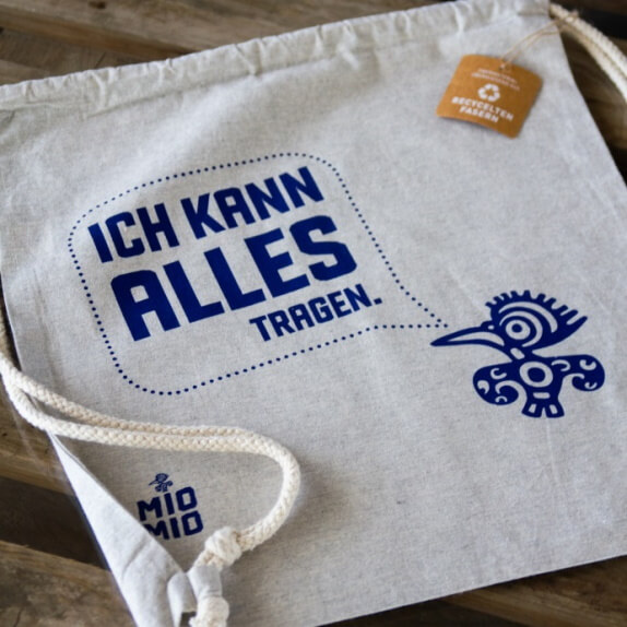 Hellgrauer Mio Mio Baumwollbeutel auf Holzboden mit blauem Logo und Schriftzug: Ich kann alles Tragen