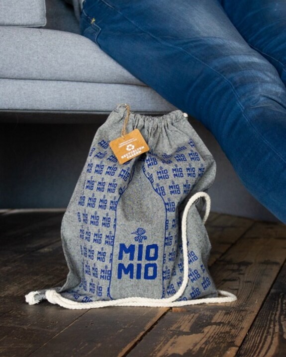 Dunkelgrauer Mio Mio Baumwollbeutel mit blauem Logo und weißen Bändern auf Holzboden vor grauem Sofa